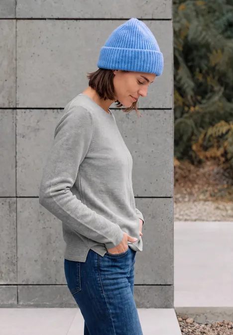 Frau mit blauem Beanie und grauem Pullover steht seitlich vor einer Betonwand.