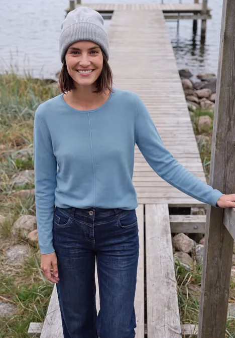 Jeune femme souriante avec bonnet gris et pull bleu debout sur une jetée au bord de l'eau.