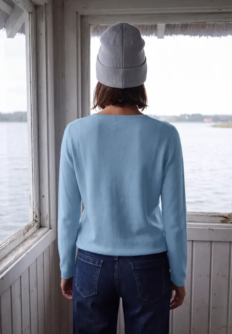 Une personne vue de dos en pull bleu clair, jean et bonnet gris sur une terrasse en bois au bord de l’eau.