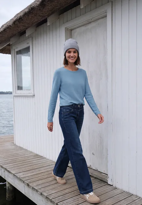 Une femme en bonnet, pull bleu clair et jean marche sur un ponton en bois devant une maisonnette blanche.