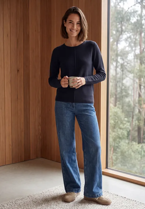 Femme souriante en pull et jean tient une tasse dans un intérieur en bois avec vue sur la forêt.