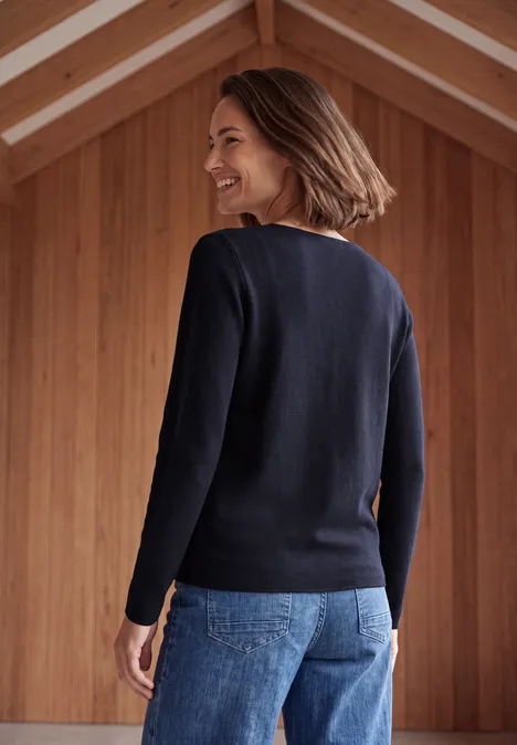 Femme de dos dans un intérieur en bois, en pull bleu foncé et jean, sourit en tournant la tête.