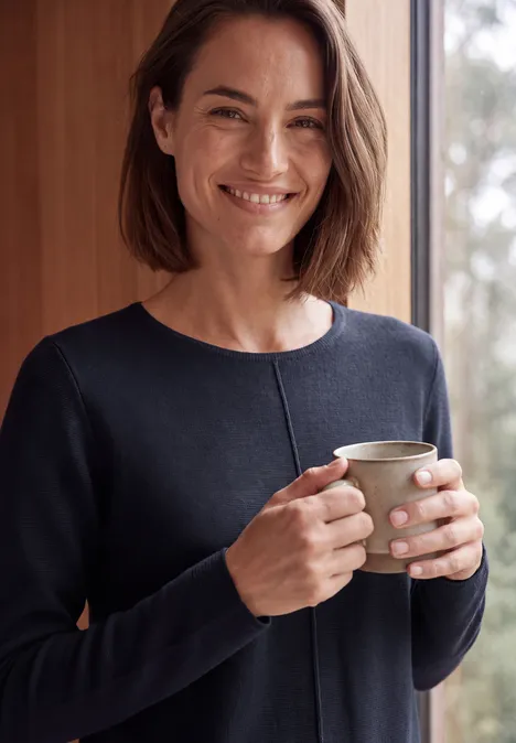 Femme en pull bleu foncé souriante tenant une tasse devant un fond lumineux.