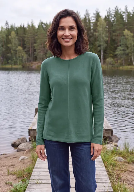Trui met ronde hals en structuur jewel green melange Eine lächelnde Frau in grünem Pullover und Jeans steht auf einem Steg am See, umgeben von Wäldern.