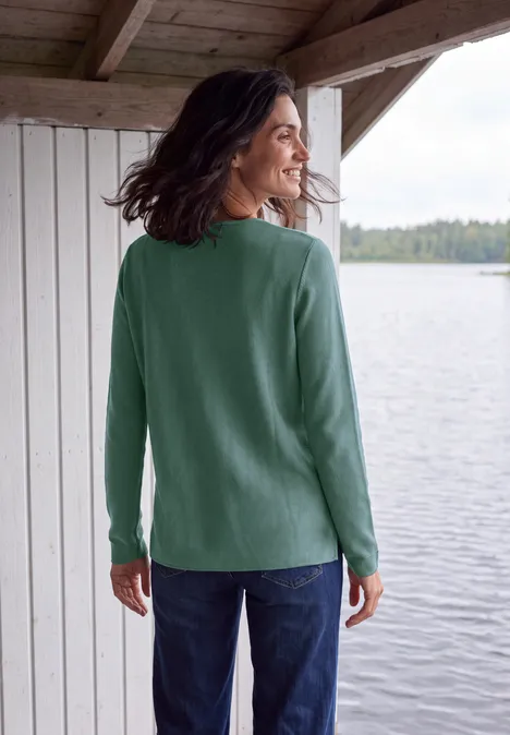 Trui met ronde hals en structuur jewel green melange Frau im grünen Pullover steht auf einer überdachten Terrasse und blickt auf einen See.