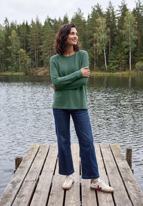 Trui met ronde hals en structuur jewel green melange Frau im grünen Pullover und Jeans steht auf einem Steg an einem ruhigen See mit Wald im Hintergrund.