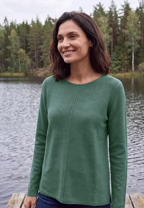 Trui met ronde hals en structuur jewel green melange Eine lächelnde Frau trägt einen grünen Pullover am Ufer eines Sees mit Wald im Hintergrund.