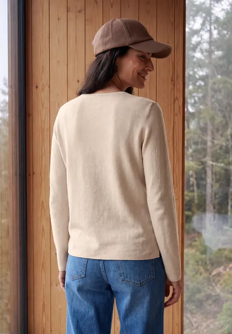 Eine Frau in beigem Pullover, blauen Jeans und brauner Cap steht im Holzzimmer vor Waldfenster.