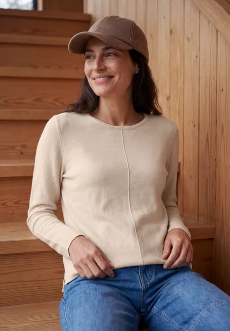 Frau mit brauner Mütze, beigefarbenem Pullover und Jeans sitzt lächelnd auf einer Holztreppe.