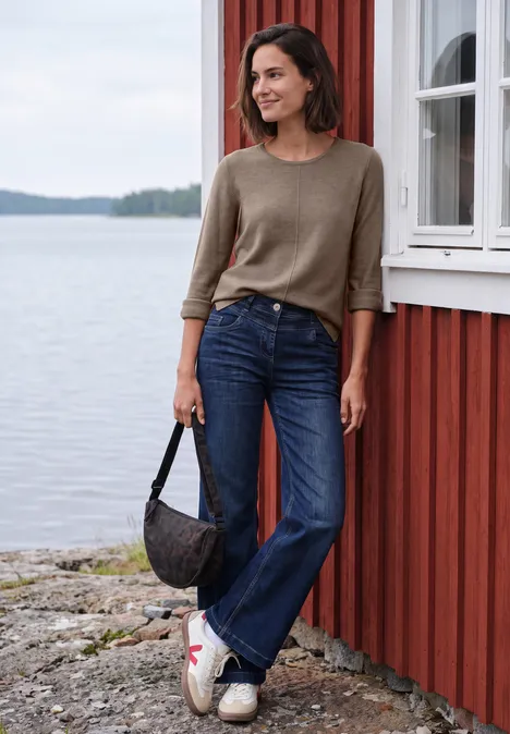 Une femme sourit, debout au bord du lac devant une maison rouge, en jean et pull, tenant un sac.