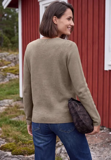 Femme en pull marron et jean, souriante de profil, tenant une petite pochette, devant une maison en bois rouge.