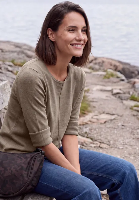 Une femme souriante est assise au bord de l’eau sur des rochers, l’air détendu.