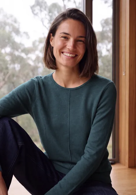Langarm Pullover mit Rundhals und Struktur dull teal blue melange Eine lächelnde Frau mit mittellangen dunklen Haaren trägt grünen Pullover und sitzt vor einem Fenster.