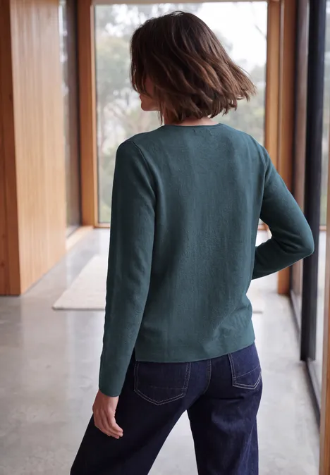 Langarm Pullover mit Rundhals und Struktur dull teal blue melange Frau steht von hinten in grünem Pullover und blauen Jeans vor großen Fenstern.