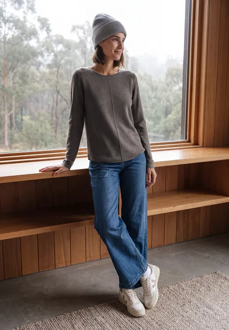 Langarm Pullover mit Rundhals und Struktur mushroom taupe melange Frau in grauem Pullover und Jeans lehnt entspannt am Fenster aus Holz, trägt Mütze und Sneakers.