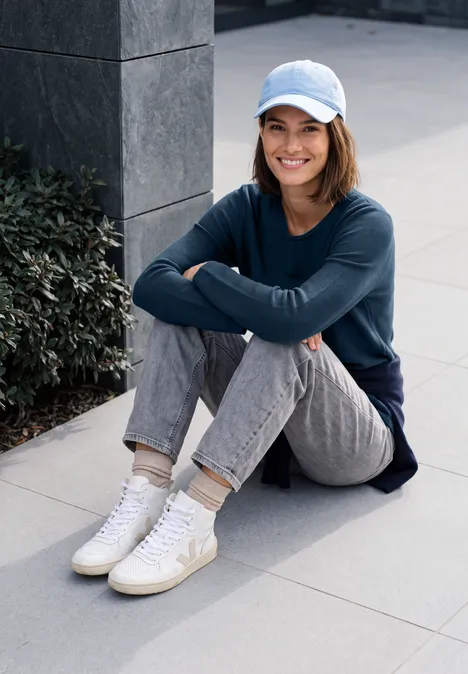 Frau mit Cap sitzt lächelnd im Freien, verschränkt die Arme auf den Knien, trägt Jeans, Pullover und Sneaker.