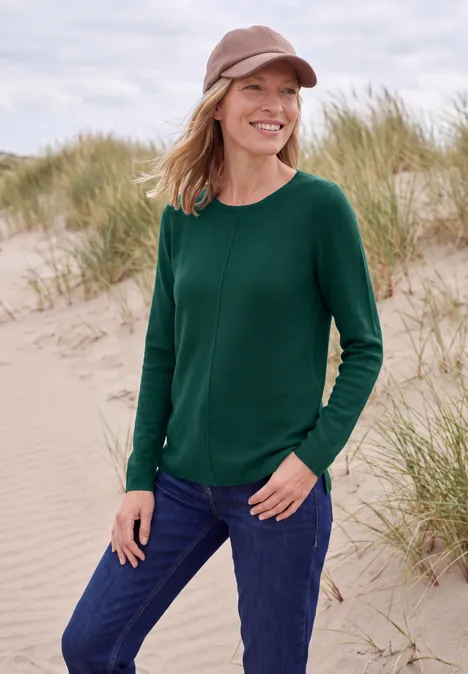 Lächelnde Frau in grünem Pullover und Mütze steht am Strand vor Sanddünen.