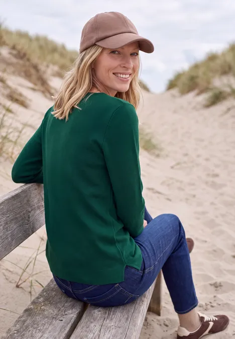 Frau im grünen Pullover und Jeans sitzt am Strand auf einem Geländer, trägt Cap und lächelt zur Seite.