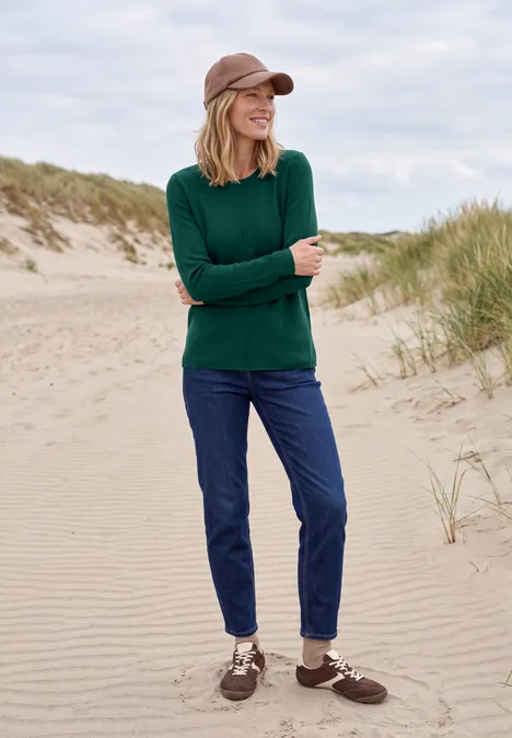 Frau in grünem Pullover, Jeans und Cap steht entspannt auf einer sandigen Düne mit Grasbüscheln.