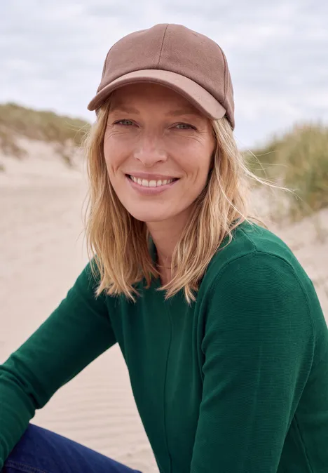 Eine lächelnde Frau mit Baseballcap und grünem Pullover sitzt am Strand, im Hintergrund Sanddünen.