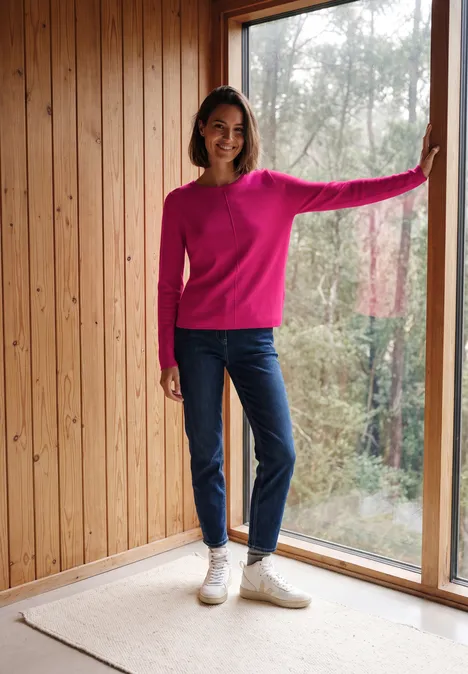Eine Frau in pinkem Pullover und Jeans steht an großer Fensterfront.