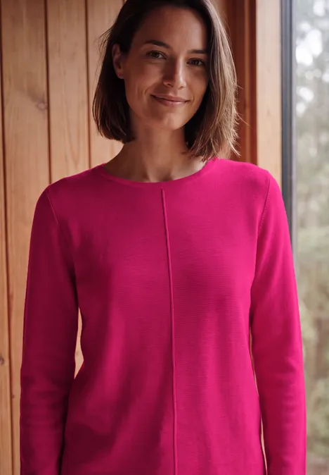 Eine lächelnde Frau in einem pinken Pullover steht in einem Raum mit Holzwänden.