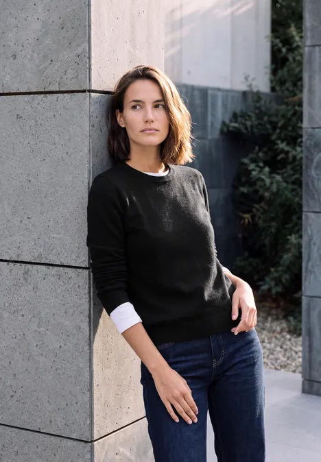 Pullover in Unifarbe mit Rundhals Black Eine Frau lehnt an einer hellen Steinwand, trägt schwarzen Pullover, Jeans und steht im Außenbereich neben Pflanzen.