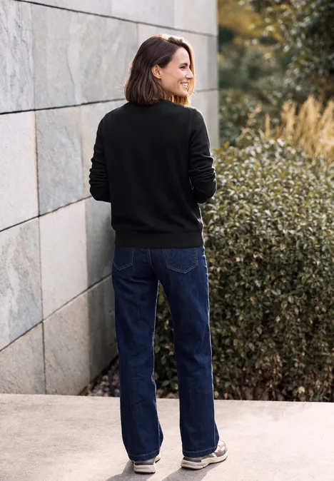 Pullover in Unifarbe mit Rundhals Black Frau im schwarzen Pullover und blauer Jeans steht mit dem Rücken zur Kamera vor Mauer und Büschen.