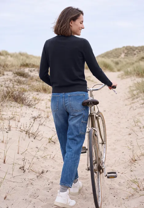 Frau schiebt Fahrrad in Jeans, schwarzem Pullover und weißen Sneakers über Dünenpfad am Strand