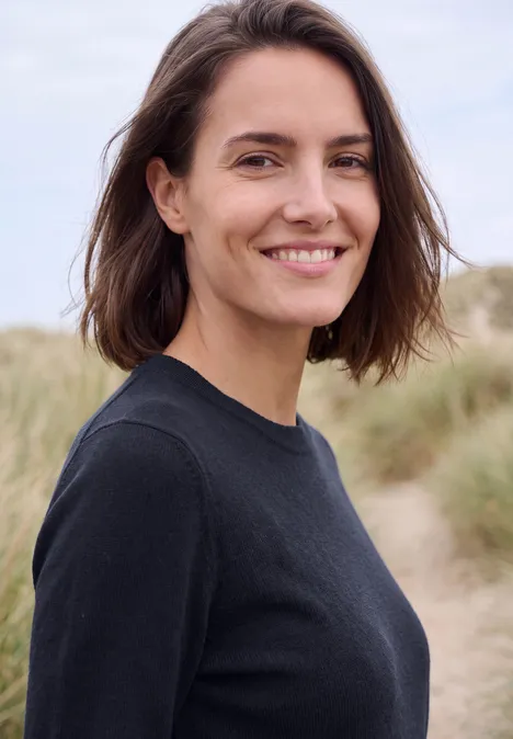 Eine lächelnde Frau mit braunen Haaren in schwarzem Pullover vor Dünenkulisse.