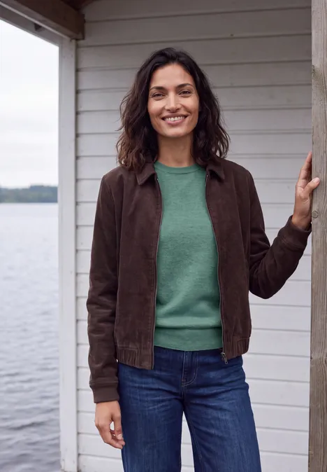 Pull uni à col rond jewel green melange Femme souriante en pull vert, veste brune et jean, devant une cabane en bois face à l'eau.