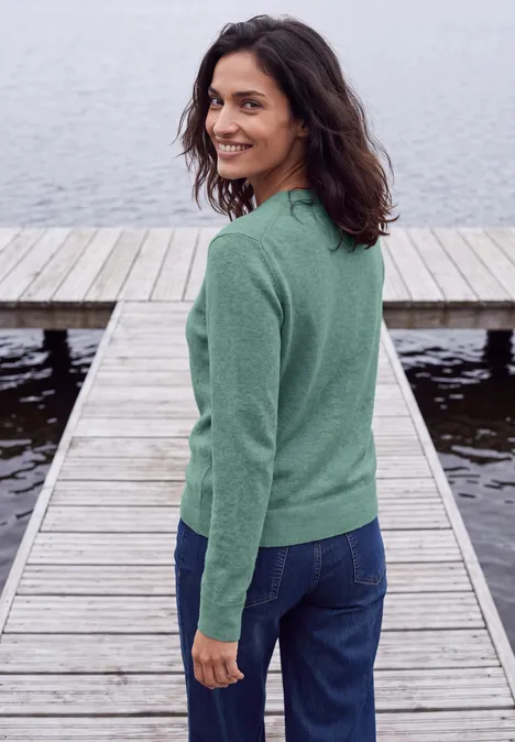 Pull uni à col rond jewel green melange Une femme en pull vert et jean se tient sur une jetée au bord de l'eau, souriant par-dessus son épaule.