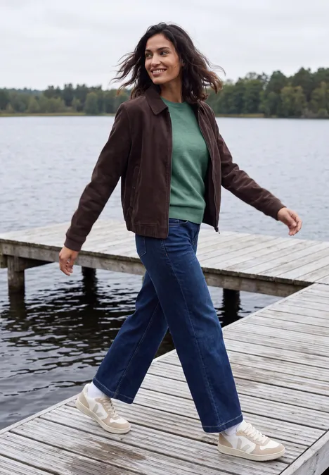 Pull uni à col rond jewel green melange Femme en pull vert et veste marron marche en souriant sur une jetée au bord d'un lac.