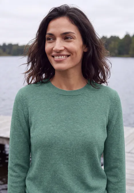 Pull uni à col rond jewel green melange Une femme souriante en pull vert devant un paysage lacustre.