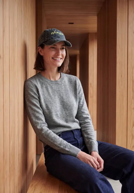 Pull uni à col rond sporty grey melange Une femme souriante assise dans une structure en bois porte un pull gris, un pantalon bleu et une casquette.