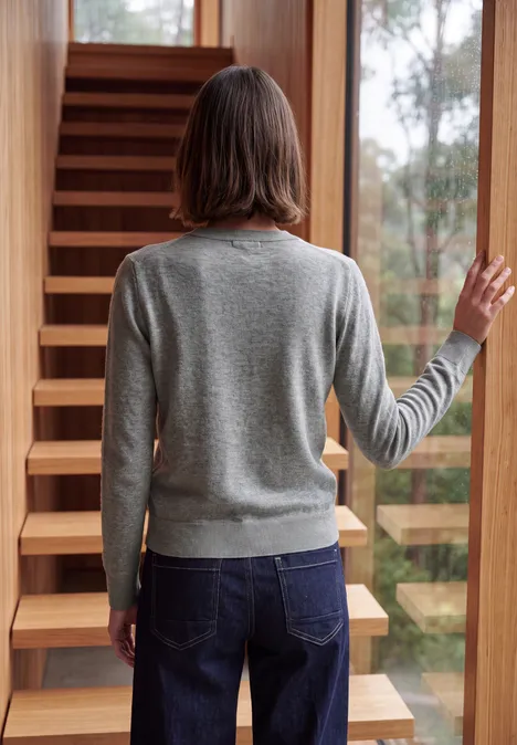 Pull uni à col rond sporty grey melange Une personne en pull gris et jean, de dos, devant un escalier en bois et une fenêtre de verre.