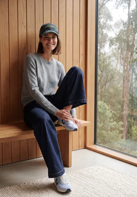 Pull uni à col rond sporty grey melange Une jeune femme en casquette, pull et jeans, souriante, assise sur un banc en bois près d'une fenêtre donnant sur la forêt.