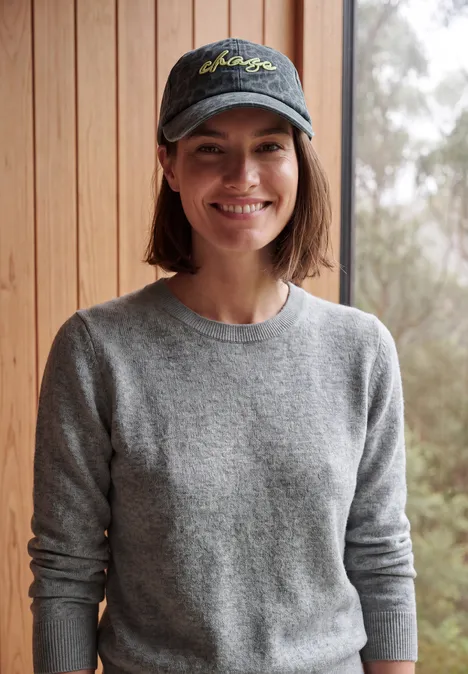 Pull uni à col rond sporty grey melange Une personne souriante en pull gris et casquette devant un mur en bois, avec des arbres en arrière-plan.