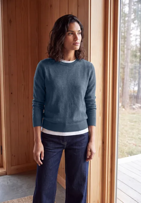 Pullover in Unifarbe mit Rundhals cotton blue melange Frau in blauem Pullover und Jeans steht in einem hellen Holzraum und blickt nach draußen.