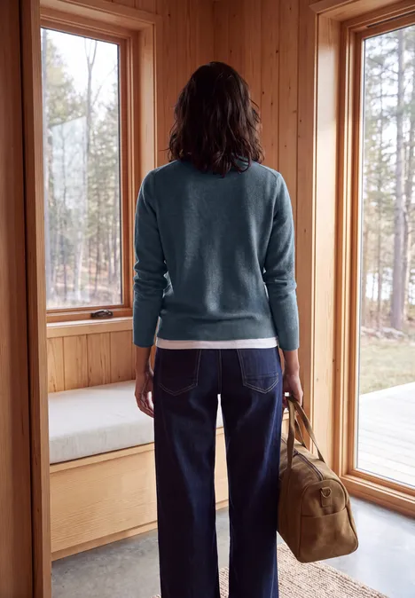 Pullover in Unifarbe mit Rundhals cotton blue melange Eine Frau im Pullover und Jeans steht in einer Holzhütte und hält eine Tasche, während sie aus dem Fenster schaut.