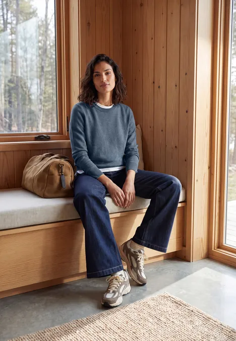 Pullover in Unifarbe mit Rundhals cotton blue melange Eine Frau in Strickpullover, Jeans und Sneakers sitzt entspannt auf einer Holzbank vor großem Fenster.