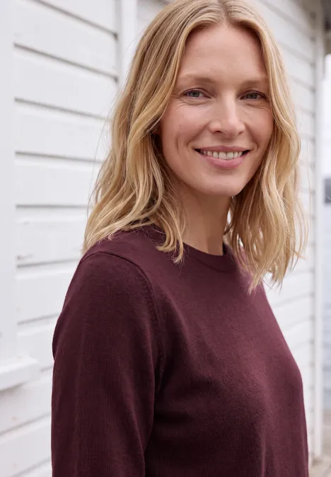 Eine lächelnde blonde Frau in einem weinroten Pullover vor einer weißen Wand.