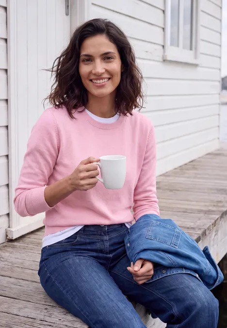 Pull uni à col rond mellow rose melange Une femme souriante en pull rose et jean, assise sur une marche en bois, tenant une tasse blanche.