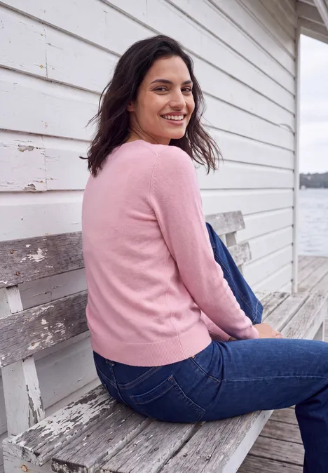 Pull uni à col rond mellow rose melange Femme en pull rose et jean assise sur un banc en bois près de l'eau, souriante en se retournant.