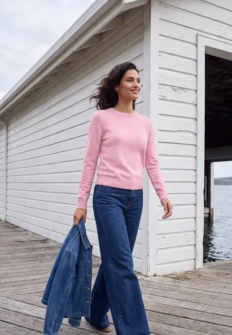 Pull uni à col rond mellow rose melange Femme en pull rose et jean tenant une veste en denim sur une passerelle près d’un hangar à bateaux.