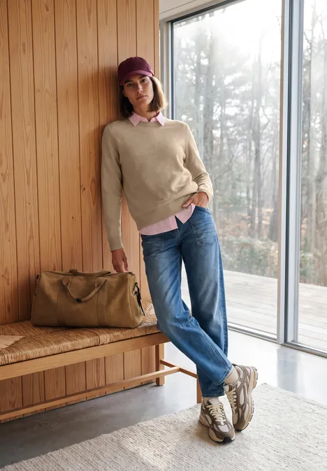 Pull uni à col rond cashew beige melange Eine Frau in Pullover, Jeans und Cap lehnt entspannt an einer Holzwand neben einer Reisetasche.