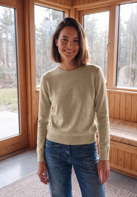 Pull uni à col rond cashew beige melange Eine lächelnde Frau in beigem Pullover und Jeans steht in einem holzverkleideten Raum vor großen Fenstern