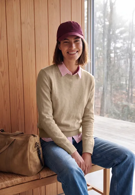 Pull uni à col rond cashew beige melange Lächelnde Person in Jeans, Pullover und Mütze sitzt mit Tasche auf Holzbank vor Hütte im Wald.