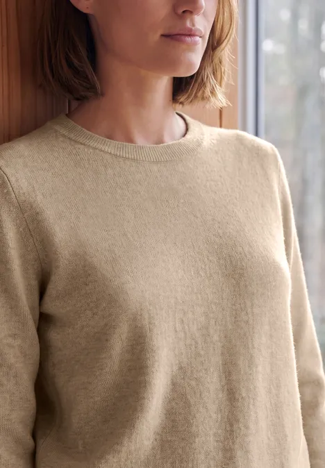 Pull uni à col rond cashew beige melange Naheinstellung einer Person in beigem Pullover vor einem Fenster.