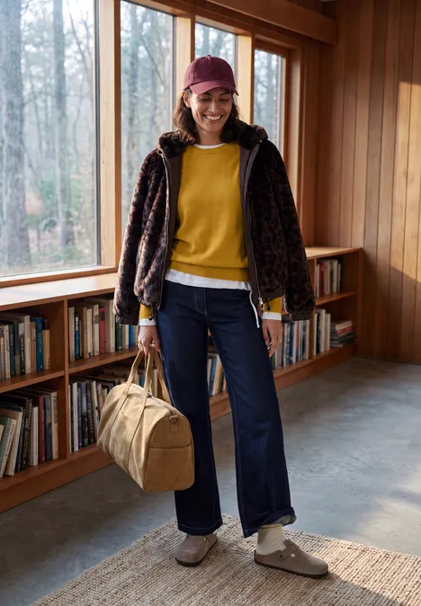 Person in gelbem Pullover, Jeans, gemusterter Jacke und Beanie hält Reisetasche in Holzraum mit Bücherregal